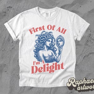 May include: A white short-sleeve t-shirt featuring a blue graphic of Medusa holding a hand mirror. The text First Of All is printed in red above the graphic, and the text I'm A Delight is printed in red below the graphic. The shirt has a crew neck.