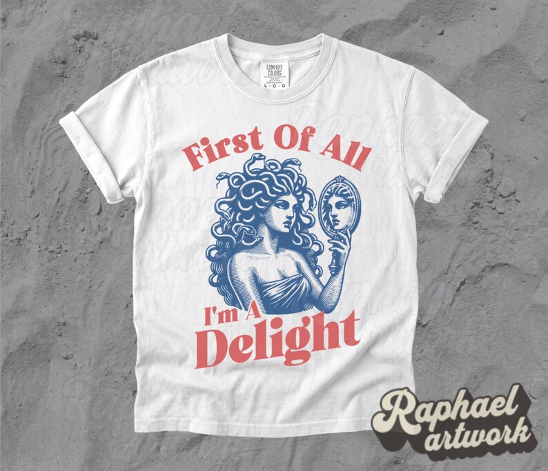 May include: A white short-sleeve t-shirt featuring a blue graphic of Medusa holding a hand mirror. The text First Of All is printed in red above the graphic, and the text I'm A Delight is printed in red below the graphic. The shirt has a crew neck.