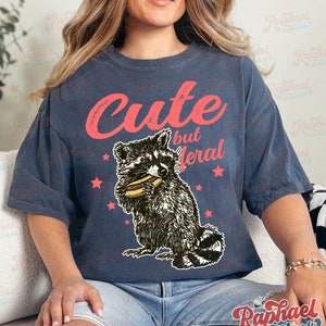 May include: Dark blue t-shirt with the text "Cute but Feral" in red script. The shirt features a graphic of a raccoon eating a hot dog, surrounded by red stars. The shirt is a relaxed fit.