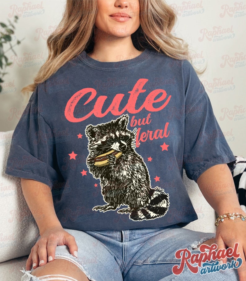 May include: Dark blue t-shirt with the text "Cute but Feral" in red script. The shirt features a graphic of a raccoon eating a hot dog, surrounded by red stars. The shirt is a relaxed fit.