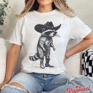 May include: A white t-shirt with a black and white illustration of a raccoon wearing a cowboy hat and boots.