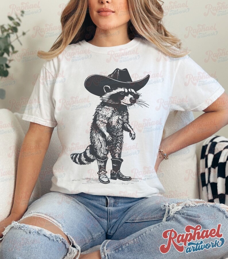 May include: A white t-shirt with a black and white illustration of a raccoon wearing a cowboy hat and boots.