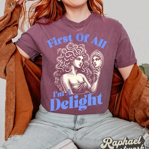 May include: A plum-colored short-sleeve t-shirt featuring a graphic print of a figure with snakes for hair looking into a hand mirror. The text "First Of All" appears in light blue above the graphic, and "I'm A Delight" appears in light blue below.