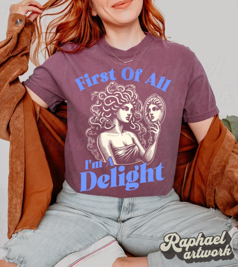 May include: A plum-colored short-sleeve t-shirt featuring a graphic print of a figure with snakes for hair looking into a hand mirror. The text "First Of All" appears in light blue above the graphic, and "I'm A Delight" appears in light blue below.