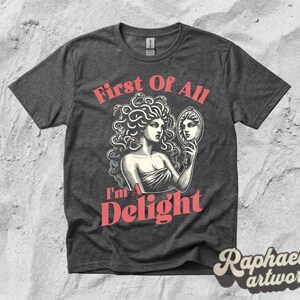 May include: A dark heather gray short-sleeve t-shirt featuring a graphic illustration of a figure with snakes for hair looking into a hand mirror. The text "First Of All I'm A Delight" is printed in a salmon pink color above and below the illustration.