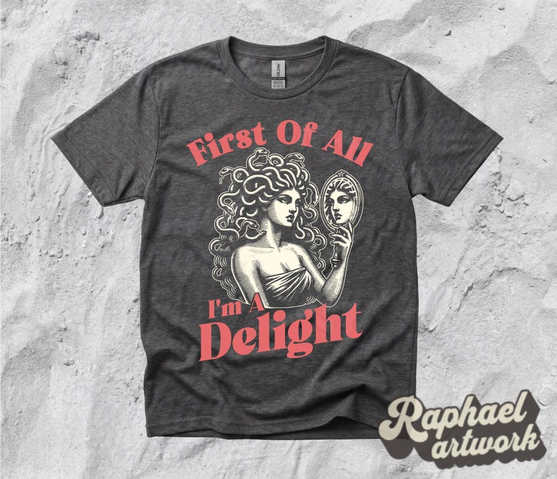 May include: A dark heather gray short-sleeve t-shirt featuring a graphic illustration of a figure with snakes for hair looking into a hand mirror. The text "First Of All I'm A Delight" is printed in a salmon pink color above and below the illustration.