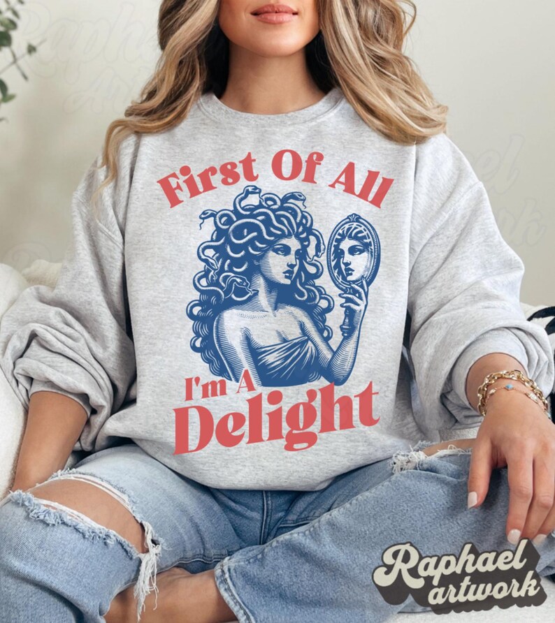 May include: A light gray crewneck sweatshirt featuring a blue graphic illustration of Medusa holding a hand mirror. The text First Of All is printed in red above the illustration, and the text I'm A Delight is printed in red below the illustration.