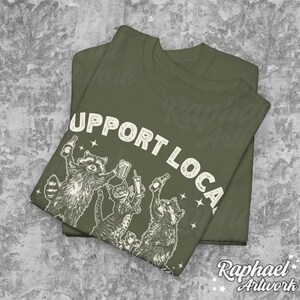 May include: A green t-shirt with the text "Support Local" printed in white. The shirt features a graphic of four raccoons drinking and holding drinks.