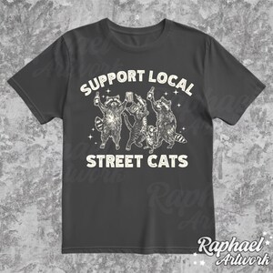 May include: A dark gray t-shirt with a white graphic of four raccoons holding drinks and the text "Support Local Street Cats".