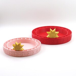 Könnte beinhalten: Zwei Adventskalender für Weihnachten, einer rot und einer rosa, mit einem goldenen Stern auf jedem. Der Stern auf dem roten Kalender sagt "X-MAS".