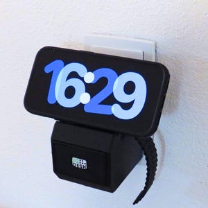 May include: A black phone charger with a black phone plugged in. The phone screen displays the time 16:29 in blue and white text. The charger has a black cord and a small white label that says "TO DESK".