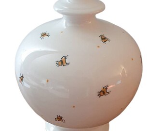 May include: A white ceramic jar with a lid, decorated with small orange and black insect illustrations. The jar has a rounded body and a pedestal base. The lid is a rounded shape with a small knob on top.