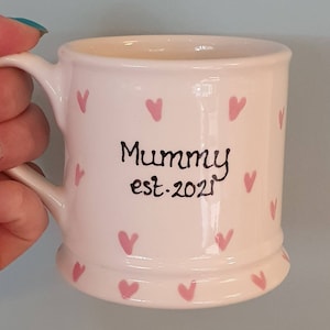 Puede incluir: Taza de cer&aacute;mica blanca con un patr&oacute;n de corazones rosas y el texto "Mummy est. 2021" en negro.