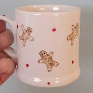 May include: White ceramic mug with a gingerbread man design. The mug has red dots and three gingerbread men with smiling faces.