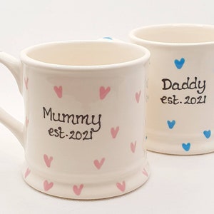 Puede incluir: Dos tazas de cer&aacute;mica blanca con corazones rosas y azules. Una taza dice "Mommy est. 2021" y la otra dice "Daddy est. 2021".