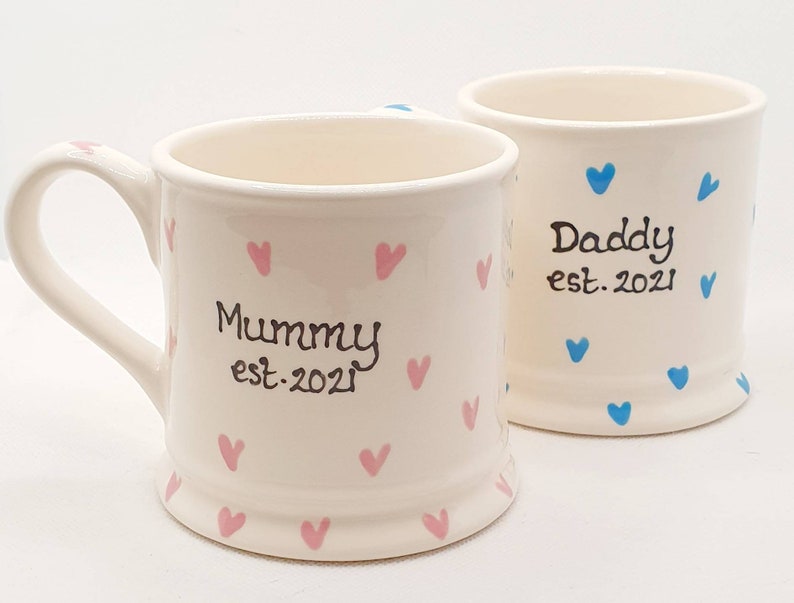 Puede incluir: Dos tazas de cer&aacute;mica blanca con corazones rosas y azules. Una taza dice "Mommy est. 2021" y la otra dice "Daddy est. 2021".