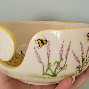 May include: Cream-colored ceramic yarn bowl with a sunflower design and a yellow rim. The bowl features painted lavender sprigs and two bees. The bowl has a curved slot to hold yarn.