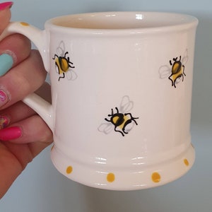 May include: White ceramic mug with a yellow and black bee design. The mug has a yellow polka dot pattern around the bottom.