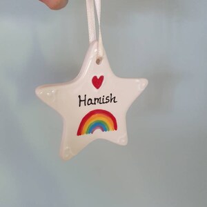 May include: White ceramic star ornament with a red heart and rainbow design. The star has the name "Hamish" written on it.