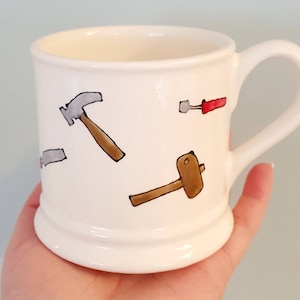 Op de afbeelding: Witte keramische mok met een handgeschilderd ontwerp van gereedschap, waaronder een hamer, schroevendraaier en hamer.
