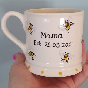 Puede incluir: Taza de cer&aacute;mica blanca con asa, con un dise&ntilde;o de abeja y el texto "Mama Est. 26.03.2021".