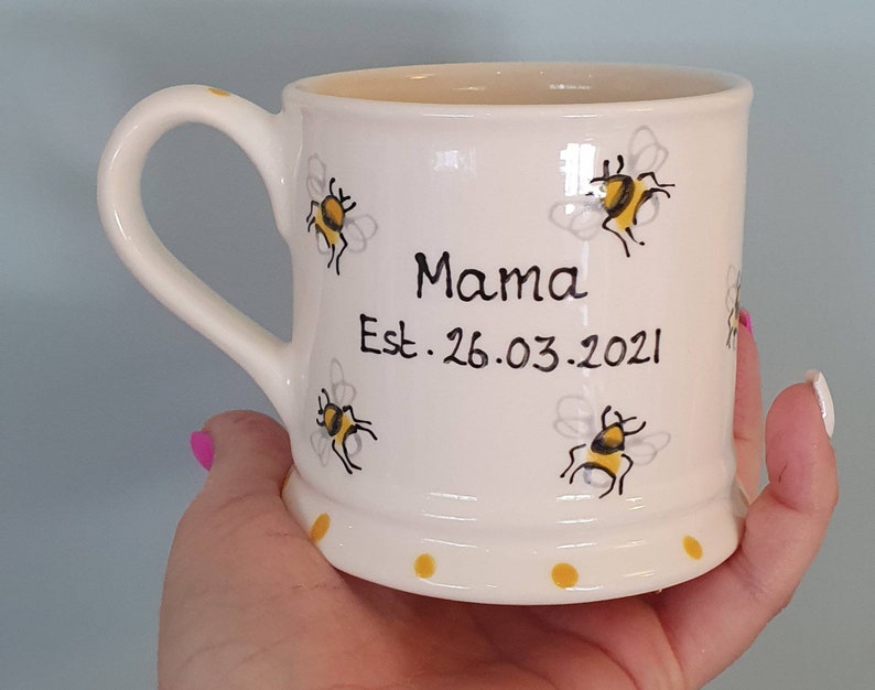 Puede incluir: Taza de cer&aacute;mica blanca con asa, con un dise&ntilde;o de abeja y el texto "Mama Est. 26.03.2021".