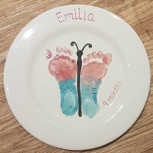 May include: A white ceramic plate featuring a butterfly design created with pink and blue handprints. The plate is personalised with the name "Emilia" and the age "9 months".