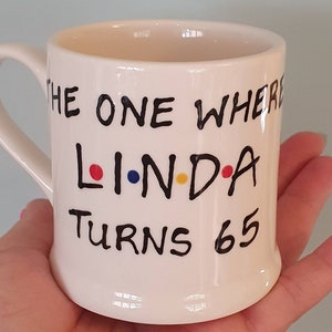 May include: A white ceramic mug with a curved handle. The mug features the text "THE ONE WHERE LINDA TURNS 65" in black lettering, with the name "LINDA" in the style of the TV show "Friends". The letters L, I, and A are colored red, blue, and yellow.