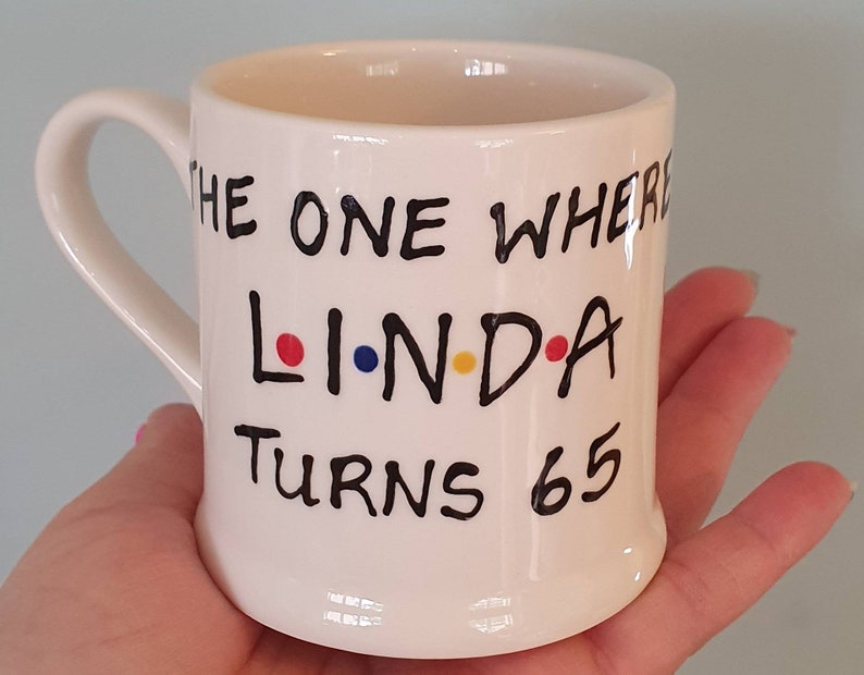 May include: A white ceramic mug with a curved handle. The mug features the text "THE ONE WHERE LINDA TURNS 65" in black lettering, with the name "LINDA" in the style of the TV show "Friends". The letters L, I, and A are colored red, blue, and yellow.