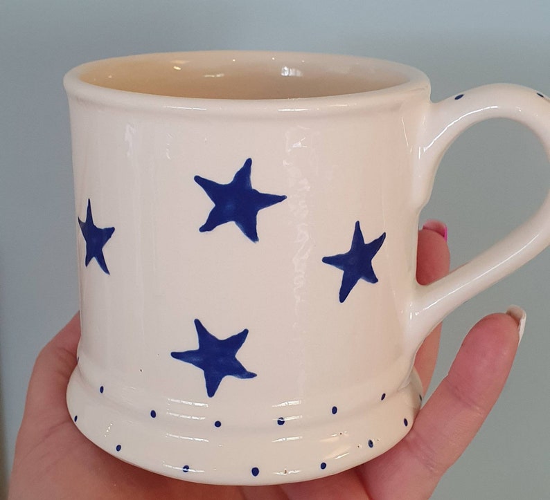 Puede incluir: Taza de cer&aacute;mica blanca con un patr&oacute;n de estrellas azules y lunares blancos alrededor de la base.