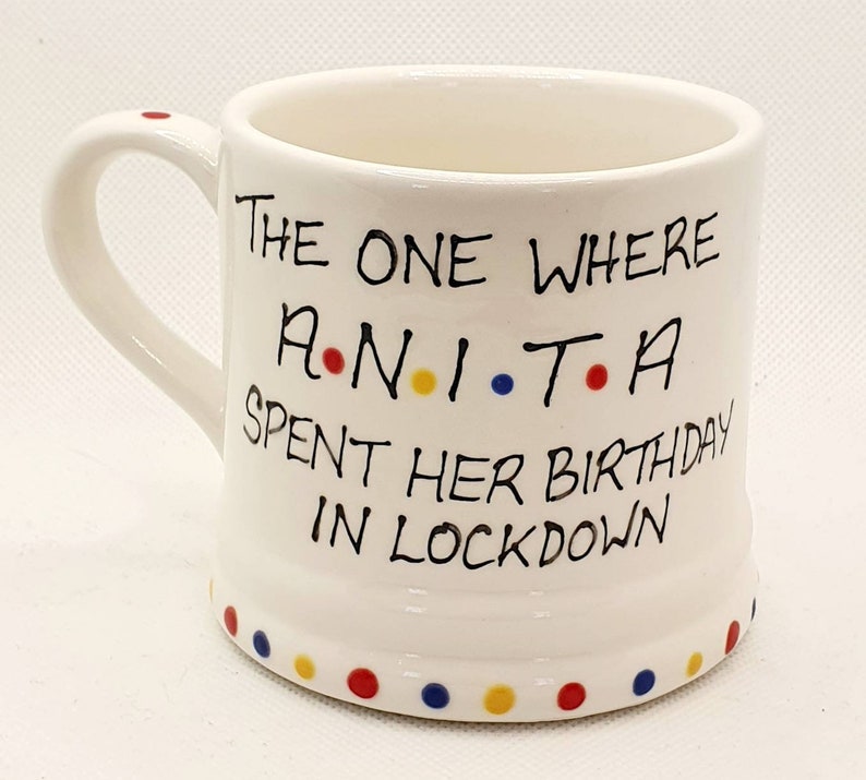 May include: White ceramic mug with a handle and the text "The One Where Anita Spent Her Birthday in Lockdown." The mug has a series of colored dots around the bottom and on the handle.