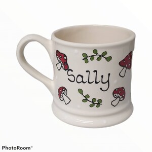 May include: White ceramic mug with a handle, featuring a red and white mushroom design, green leaves, and white polka dots. The mug has the name "Sally" written on it.
