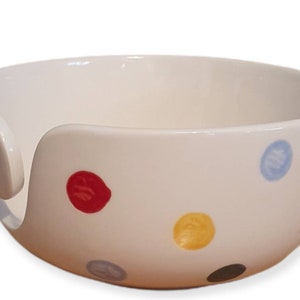 Op de afbeelding: Witte keramische garenkom met een reeks kleurrijke stippen in rood, blauw, geel, grijs en zwart. De kom heeft een gebogen opening aan de zijkant om garen vast te houden tijdens het breien of haken. Een kleine groene stip zit aan de zijkant.