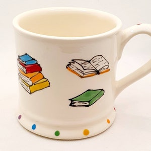 May include: White ceramic mug with a colourful illustration of books. The mug has a handle and a dotted rim.