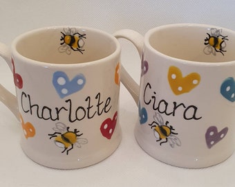 Op de afbeelding: Twee witte keramische mokken met handvatten. De mokken zijn versierd met kleurrijke harten, bijen en de namen "Charlotte" en "Ciara".