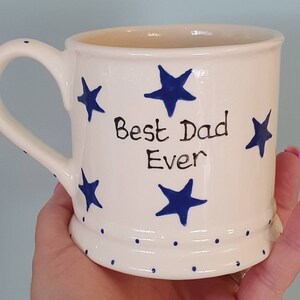 Puede incluir: Taza de cer&aacute;mica blanca con estrellas azules y el texto "Best Dad Ever" en negro. La taza tiene un borde blanco y un asa blanca.