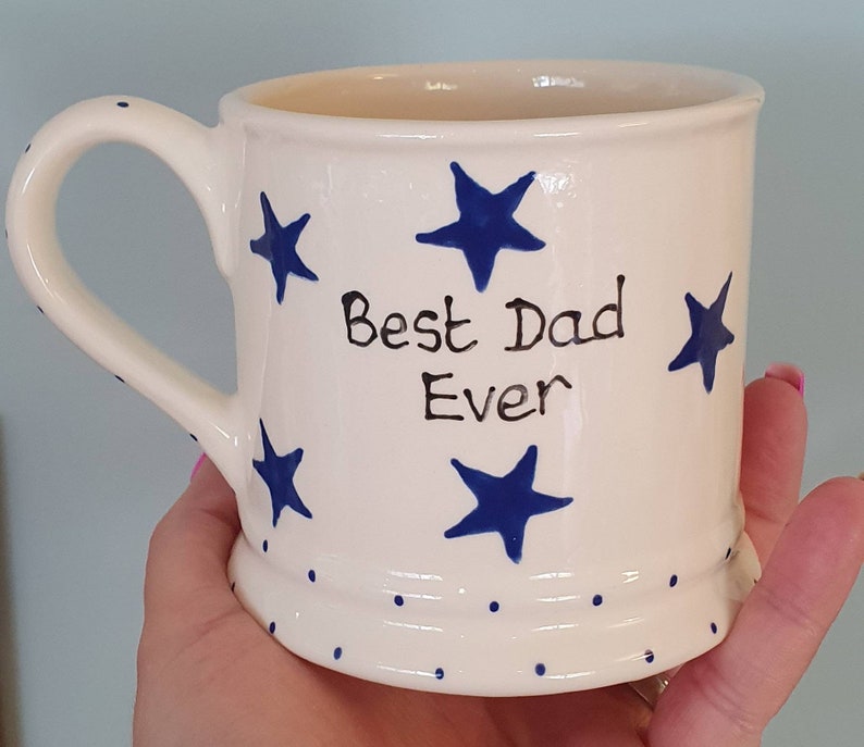 Puede incluir: Taza de cer&aacute;mica blanca con estrellas azules y el texto "Best Dad Ever" en negro. La taza tiene un borde blanco y un asa blanca.