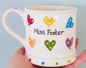 Puede incluir: Taza de cerámica blanca con asa, decorada con corazones de colores y lunares. La taza tiene el texto "Miss Foster" escrito en ella.