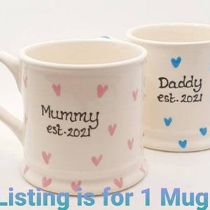 Puede incluir: Dos tazas de cer&aacute;mica blanca con corazones rosas y azules. Una taza dice "Mummy est. 2021" y la otra dice "Daddy est. 2021".