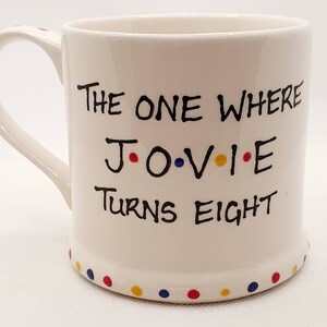 May include: White ceramic mug with the text "THE ONE WHERE J.O.V.I.E TURNS EIGHT" in black lettering. The letters J, O, V, I, and E are accented with red, blue, yellow, and red dots. The base of the mug has a row of colorful dots.