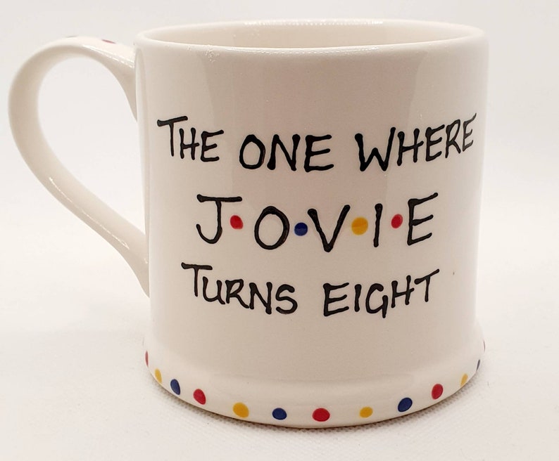 May include: White ceramic mug with the text "THE ONE WHERE J.O.V.I.E TURNS EIGHT" in black lettering. The letters J, O, V, I, and E are accented with red, blue, yellow, and red dots. The base of the mug has a row of colorful dots.