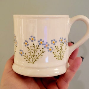 May include: White ceramic mug with a blue and yellow floral design. The mug has a wide base and a rounded handle.