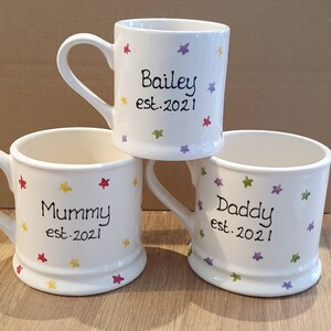 Puede incluir: Tres tazas de cer&aacute;mica blanca con dise&ntilde;os de estrellas de colores. Las tazas est&aacute;n personalizadas con los nombres "Bailey est. 2021", "Mam&aacute; est. 2021" y "Pap&aacute; est. 2021".