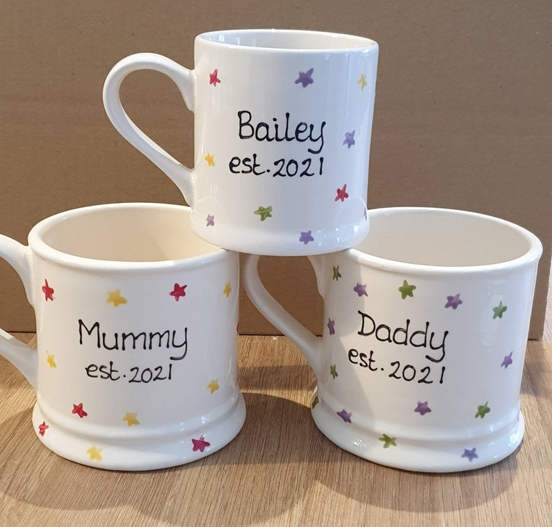 Puede incluir: Tres tazas de cer&aacute;mica blanca con dise&ntilde;os de estrellas de colores. Las tazas est&aacute;n personalizadas con los nombres "Bailey est. 2021", "Mam&aacute; est. 2021" y "Pap&aacute; est. 2021".