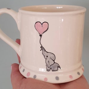 May include: A white ceramic mug with a grey and pink polka dot trim. The mug features a hand-painted design of a grey elephant holding a pink heart-shaped balloon.