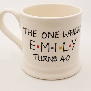 May include: Cream-colored ceramic mug with a curved handle. The mug features black text that reads "THE ONE WHERE EMILY TURNS 40" with the name in colorful dots. A birthday-themed gift.