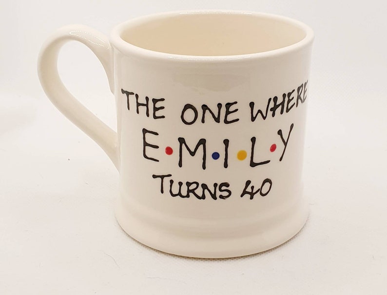 May include: Cream-colored ceramic mug with a curved handle. The mug features black text that reads "THE ONE WHERE EMILY TURNS 40" with the name in colorful dots. A birthday-themed gift.