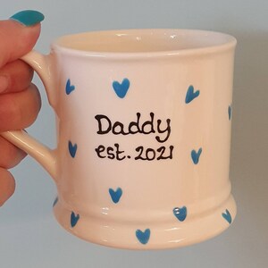 Puede incluir: Taza de cer&aacute;mica blanca con corazones azules y el texto "Daddy est. 2021" en negro.