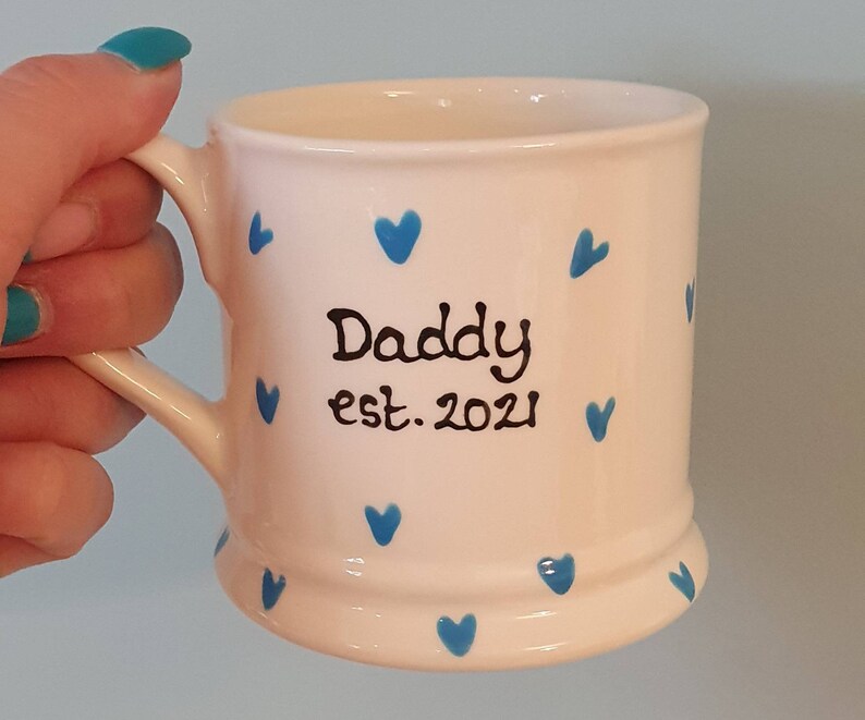 Puede incluir: Taza de cer&aacute;mica blanca con corazones azules y el texto "Daddy est. 2021" en negro.