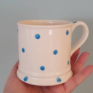 Puede incluir: Una taza de cer&aacute;mica blanca con un patr&oacute;n de lunares azules. La taza tiene un asa y una base redondeada.
